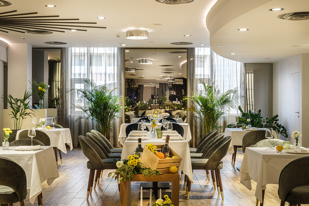 Арендовать Итальянский ресторан Umberto(Умберто). Ресторан с обеденной зоной, круглые столы с белыми скатертями, обитые стулья темного цвета, деревянный пол. Декор — живые растения и цветы. Современное освещение, включая точечные светильники и люстры. Светлые стены и окна с прозрачными шторами.