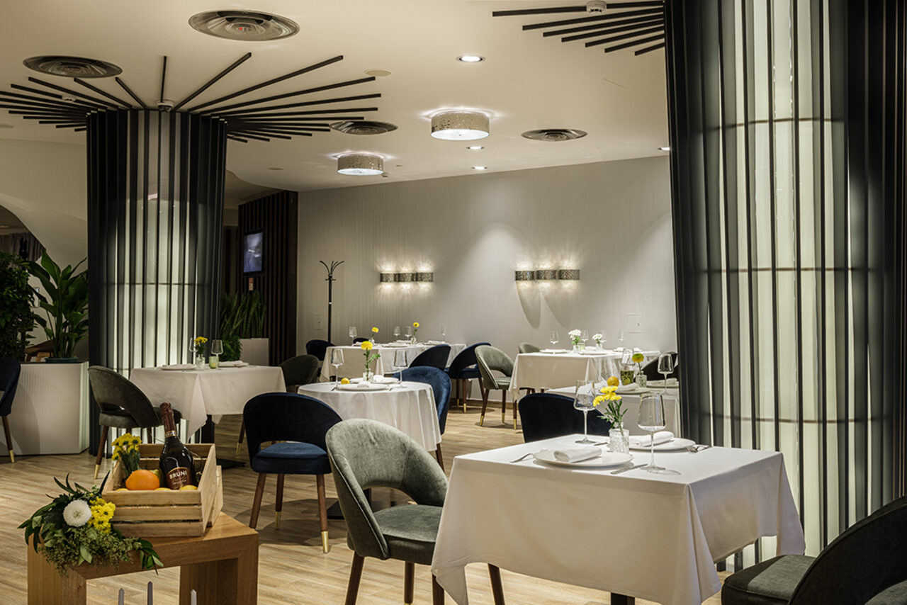 Арендовать Итальянский ресторан Umberto(Умберто). Ресторанное пространство с квадратными столами, покрытыми белыми скатертями, и темными стульями. Светлый деревянный пол, что подвешено на потолке освещение, большие колонны, декоративные элементы, растения в углу. Стены светлые с настенными светильниками.