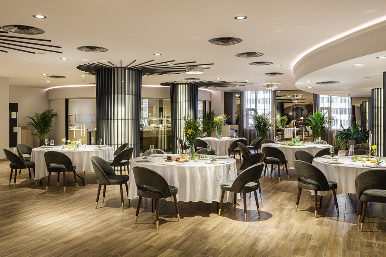 Арендовать Итальянский ресторан Umberto(Умберто). Просторное ресторанное пространство с круглыми столами, накрытыми белыми скатертями. Мягкие темные стулья, деревянный пол, круговые светильники на потолке. Расставлены декоративные растения. Светлые стены, уютное освещение.