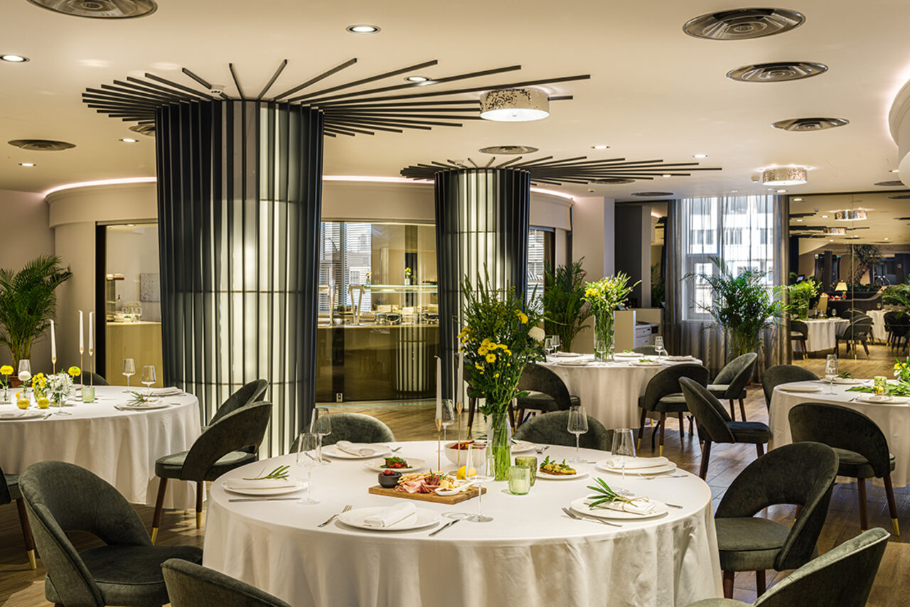 Арендовать Итальянский ресторан Umberto(Умберто). Ресторан с круглыми столами, покрытыми белыми скатертями, серые стулья, центральные колонны с декоративными элементами, деревянный пол, зеленые растения, искусственное освещение из потолочных светильников, большие окна с полупрозрачными занавесками.