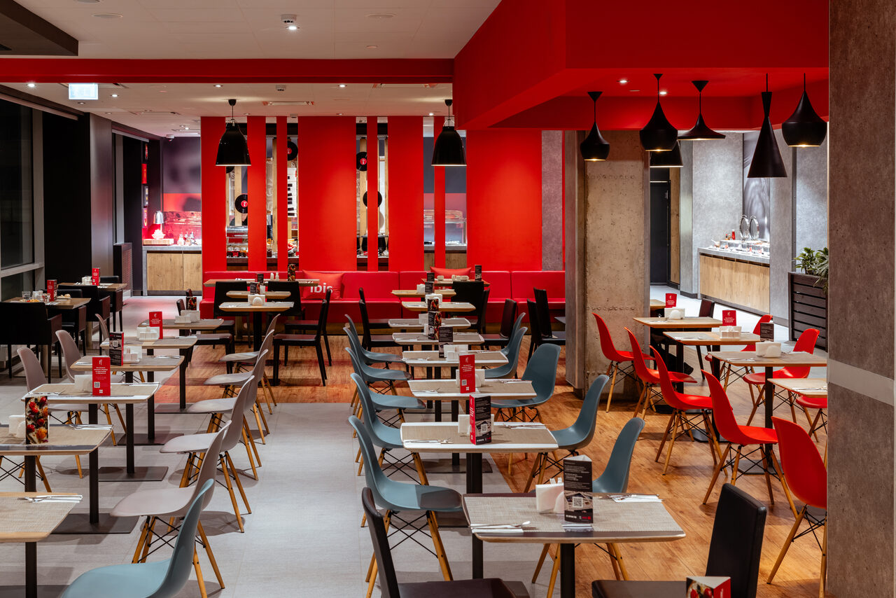 Арендовать Ресторан Ibis Kitchen(Ибис Кухня). Ресторан с открытой планировкой, множество столов и стульев; черные и красные кресла, белые столы. Яркое освещение, красные и серые стены, деревянный пол. Черные подвесные светильники, красные акценты, белый потолок.