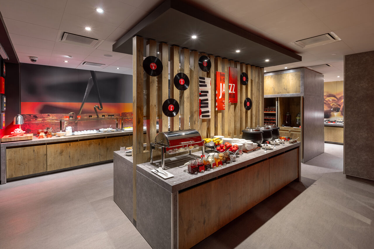 Арендовать Ресторан Ibis Kitchen(Ибис Кухня). Кейтеринговое помещение с деревянными панелями и серыми столешницами. На стене виниловые пластинки и плакат с надписью "Jazz". На столе еда и посуда. Потолочные светильники, светло-серое напольное покрытие.