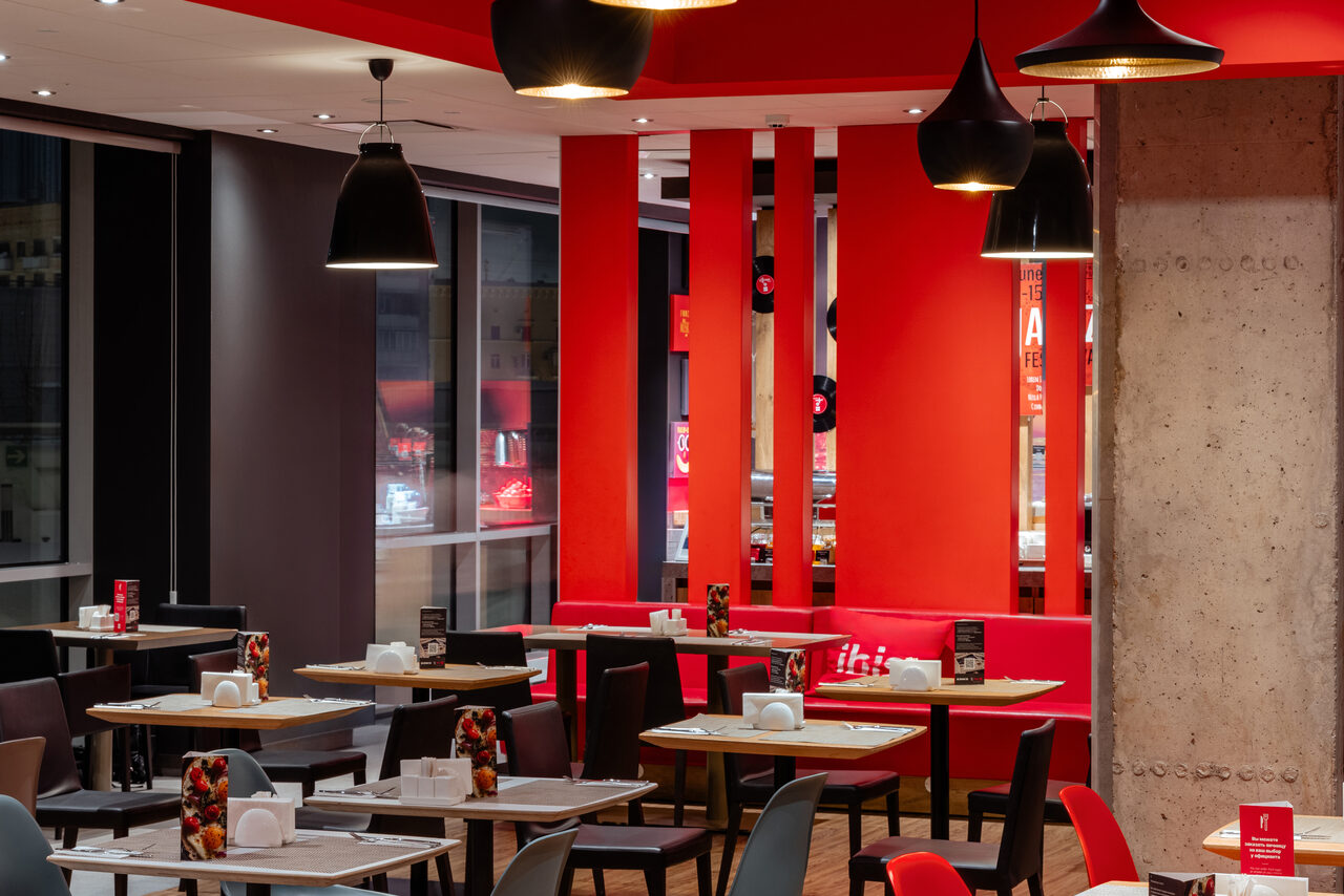 Арендовать Ресторан Ibis Kitchen(Ибис Кухня). Современное кафе с красными и черными акцентами. Столы с черными и серыми стульями, большие окна. Потолочные светильники, бетонная стена, декоративные панели, меню на столах. Яркое освещение, чистое состояние.