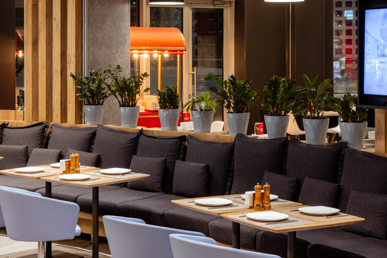 Арендовать Ресторан Ibis Kitchen(Ибис Кухня). Помещение ресторана с длинным диваном, деревянными столами и белыми стульями. На столах белая посуда и перечницы. Сзади дивана растения в серых горшках. На стене телевизор. Яркое освещение. В углу красный попкорн-аппарат.