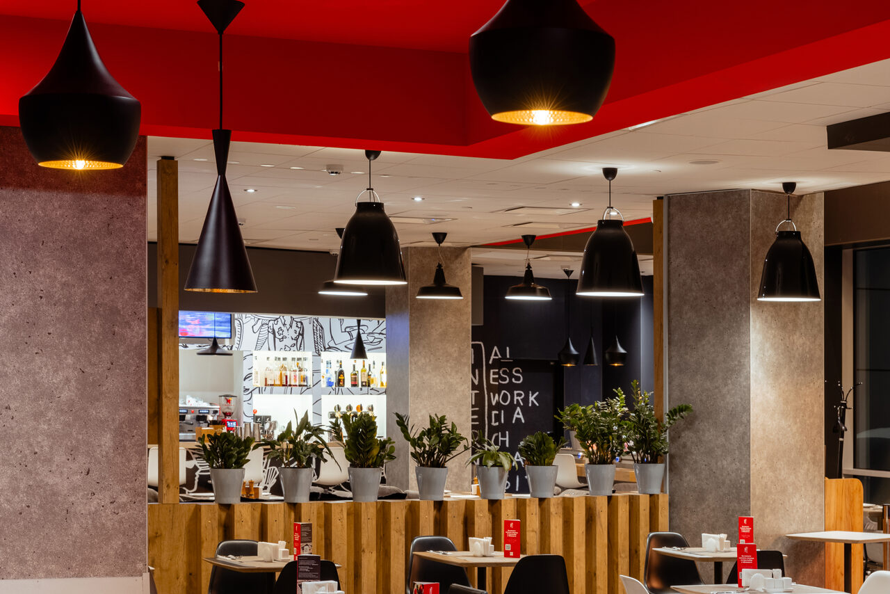Арендовать Ресторан Ibis Kitchen(Ибис Кухня). Кафе с деревянными столами и стульями, оформленными в серых и черных тонах. На столах меню в красных держателях. Потолок белый и красный, светильники черные подвесные. Присутствуют растения в белых горшках. На стене текстовое граффити.