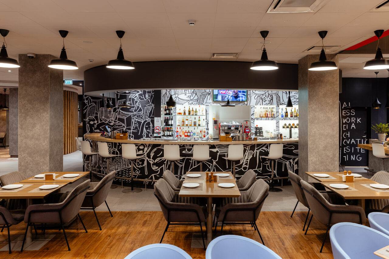 Арендовать Ресторан Ibis Kitchen(Ибис Кухня). Модное кафе с открытой планировкой. Барная стойка с высокими белыми стульями и графическим оформлением задней стенки. Деревянные обеденные столы с сервировкой и мягкими стульями. Подвесное освещение, деревянный пол, бетонные колонны.
