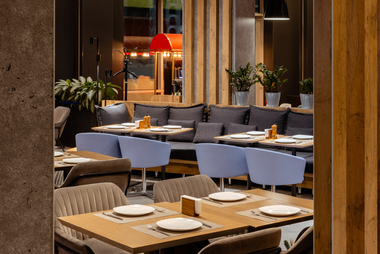 Арендовать Ресторан Ibis Kitchen(Ибис Кухня). Ресторанное пространство с деревянными столами, серыми креслами и синими диванами. На столах белая посуда. Вертикальные деревянные панели и растения в горшках. Яркое освещение, в центре зала светильники. Стены темные, полки с предметами.