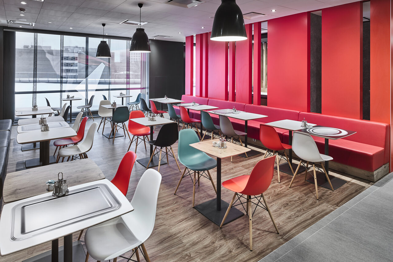 Арендовать Ресторан Ibis Kitchen(Ибис Кухня). Современное кафе с массивными окнами и видом на улицу, деревянный пол, разноцветные пластиковые стулья и квадратные столы. Красные и черные акценты, мягкие лавки вдоль стены. Потолочные светильники и яркая настенная отделка.