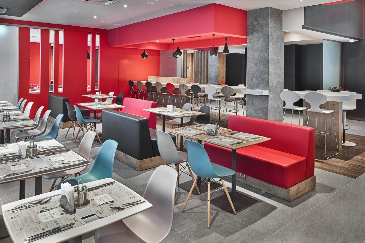 Арендовать Ресторан Ibis Kitchen(Ибис Кухня). Современное кафе с ярким интерьером: красные и серые стены, открытая планировка с множеством столов и стульев, основным цветом является красный. Зоны с диванами и барными стульями, подвесные светильники, светло-серые полы, меню на столах.