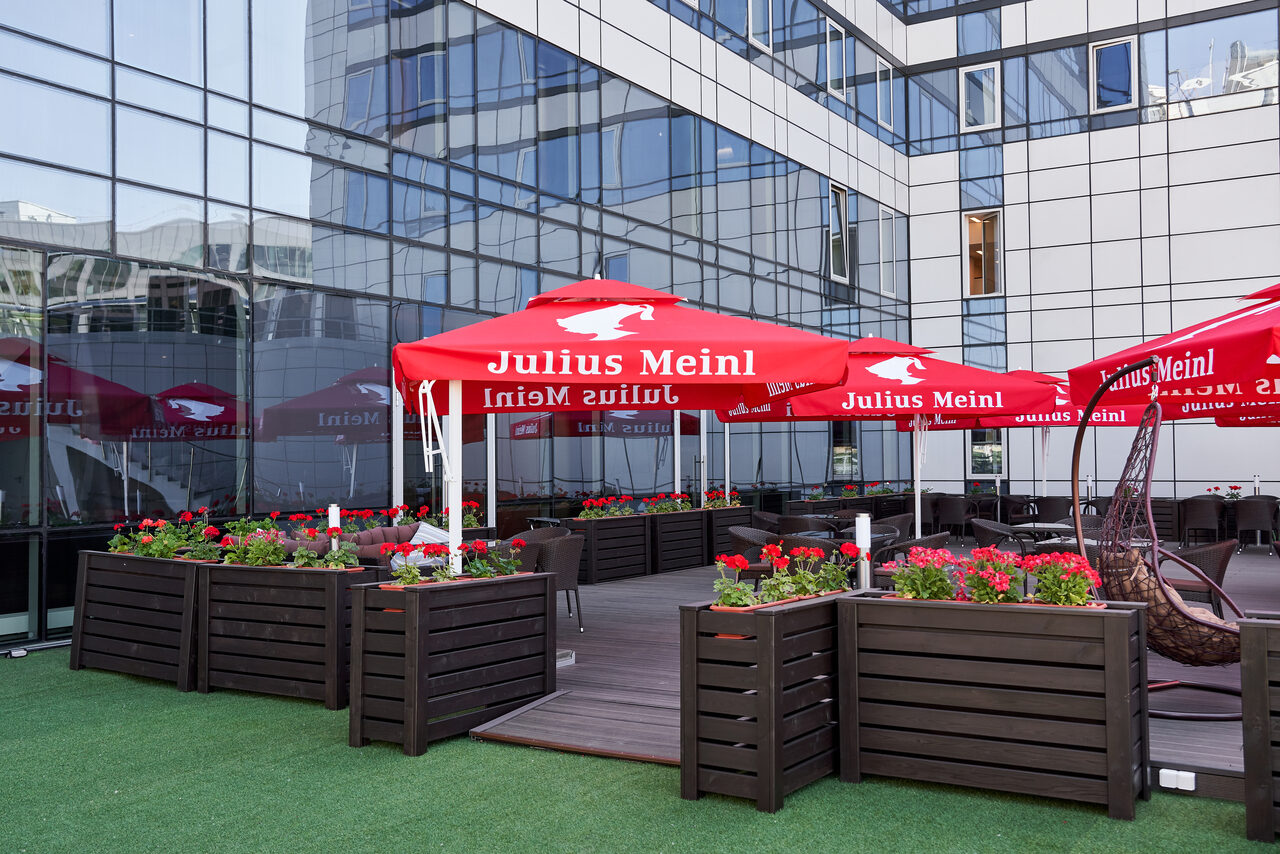 Арендовать Ресторан Ibis Kitchen(Ибис Кухня). Открытая терраса с деревянной мебелью и цветочными кадками. Над столами установлены красные зонты. Пол выстлан искусственной травой. Здание вокруг с фасадом из стекла и металла.