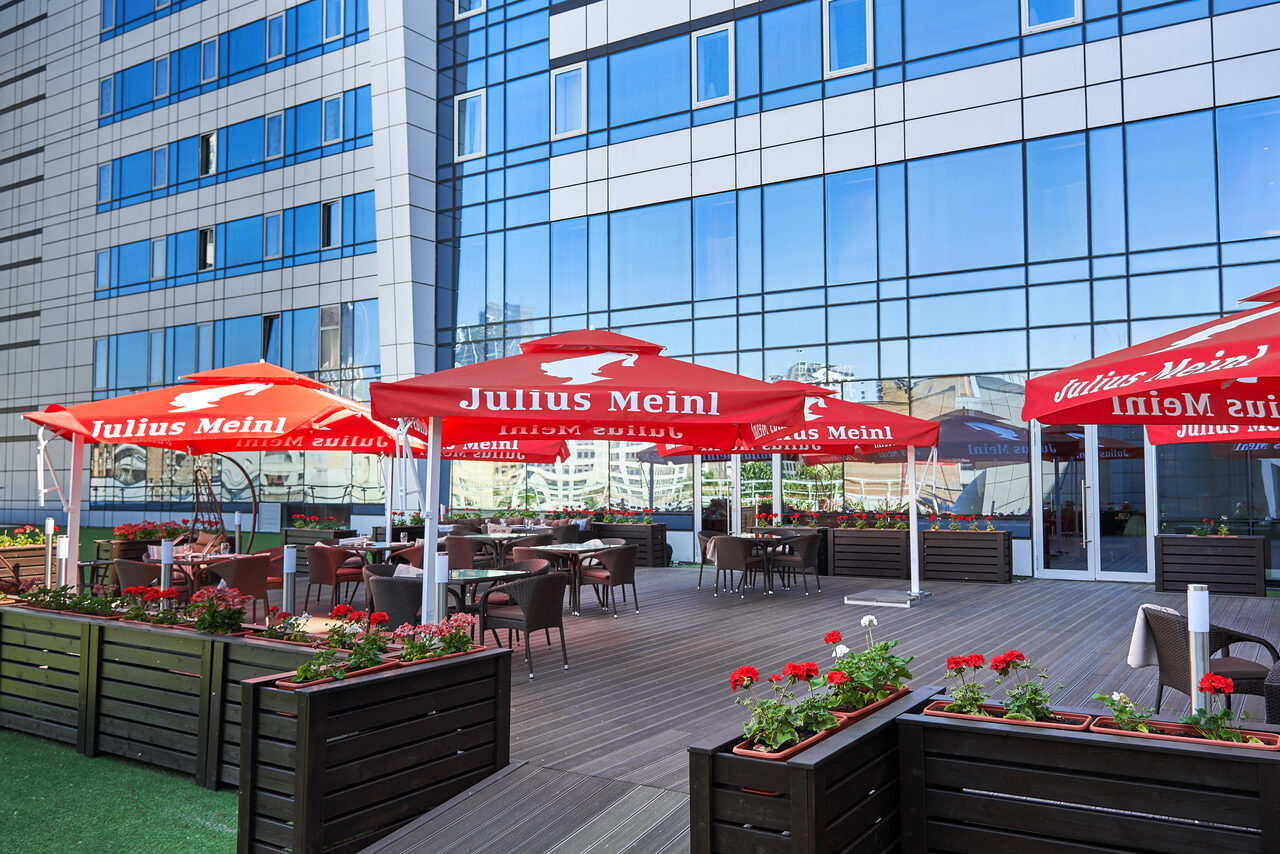 Арендовать Ресторан Ibis Kitchen(Ибис Кухня). Открытая терраса с деревянным настилом, окруженная стеклянными фасадами здания. На террасе расположены столы и стулья из темного материала. Красные зонты на стойках обеспечивают тень. Декоративные кашпо с цветами вдоль периметра.