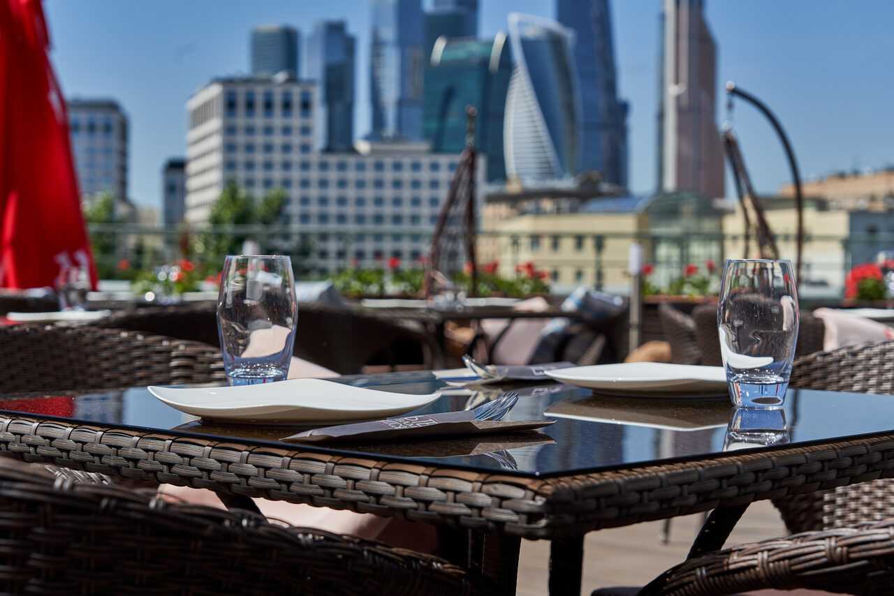 Арендовать Ресторан Ibis Kitchen(Ибис Кухня). Открытая терраса с плетеными столами и креслами, серо-коричневого цвета. На столе стеклянная поверхность, два бокала и керамическая посуда. Видны городские многоэтажки на фоне. Яркое дневное освещение.