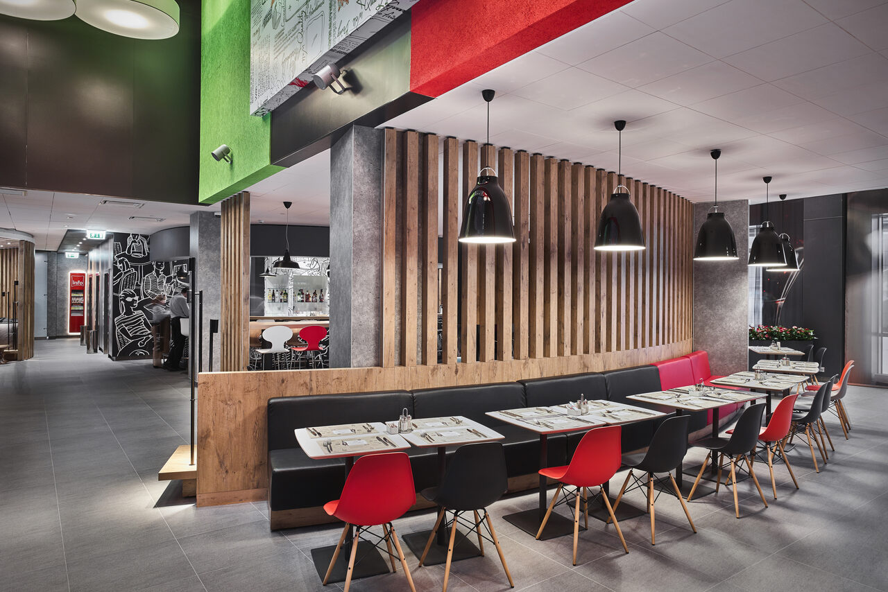 Арендовать Ресторан Ibis Kitchen(Ибис Кухня). Современное кафе с открытой планировкой; столы с черными и красными стульями; подвесные светильники; деревянные и серые элементы декора; граффити на стенах; плитка на полу; яркие акценты на потолке.