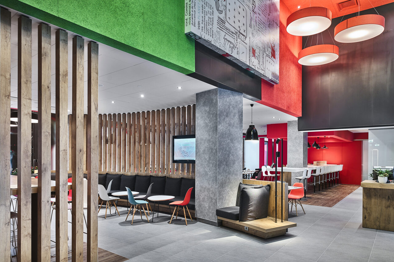 Арендовать Ресторан Ibis Kitchen(Ибис Кухня). Современное помещение с яркими цветами и открытой планировкой. Деревянный и бетонный декор. Зона отдыха с диванами и стульями рядом с экраном. Барная стойка с высокими стульями. Подвесные светильники. Белые и красные акценты.