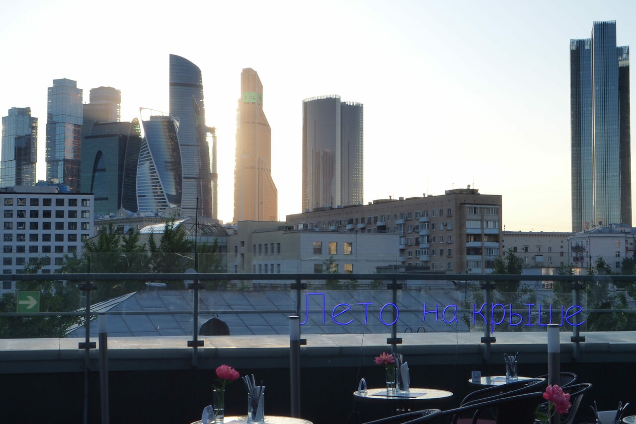 Арендовать Ресторан Ibis Kitchen(Ибис Кухня). ```plaintext
Терраса на крыше с круглыми столиками и стульями, огражденная стеклянными перилами. На столах вазы с розовыми цветами. На фоне высотные здания. Видно неоновую надпись «Лето на крыше».
```