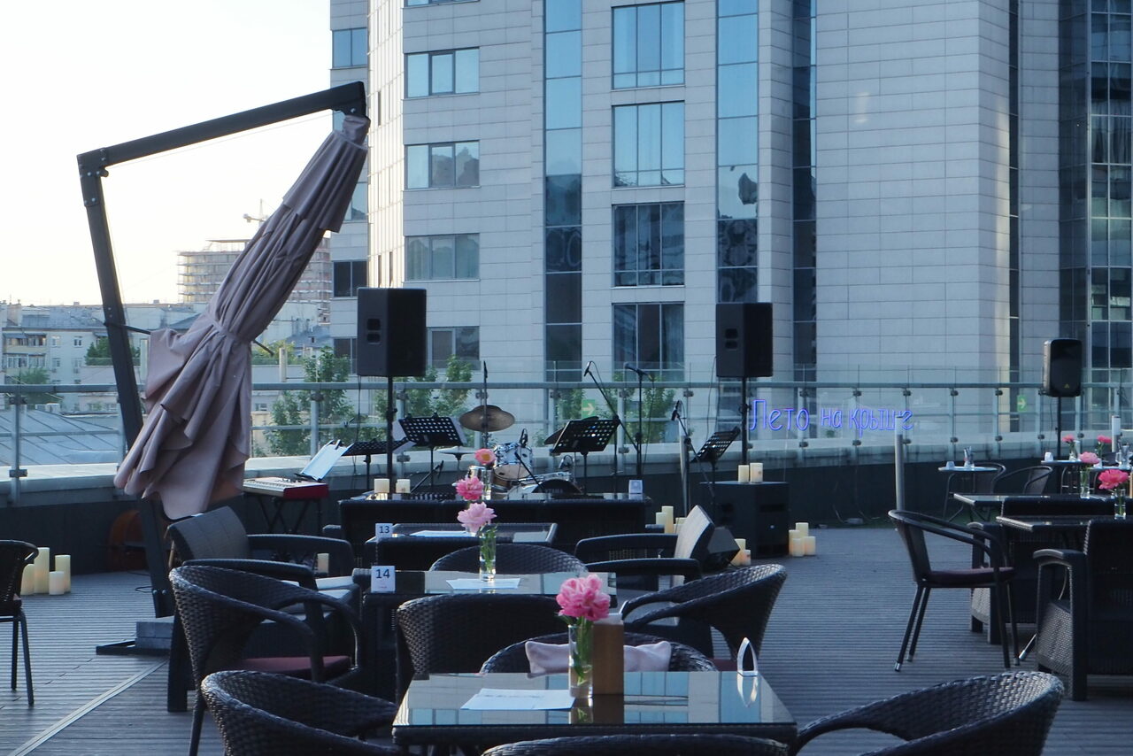 Арендовать Ресторан Ibis Kitchen(Ибис Кухня). Открытая терраса с видом на городские здания, столы и кресла из ротанга, стеклянные столешницы с розовыми цветами, звуковое оборудование для выступлений, барабанная установка, свечи вдоль перил, большие складывающиеся зонты.
