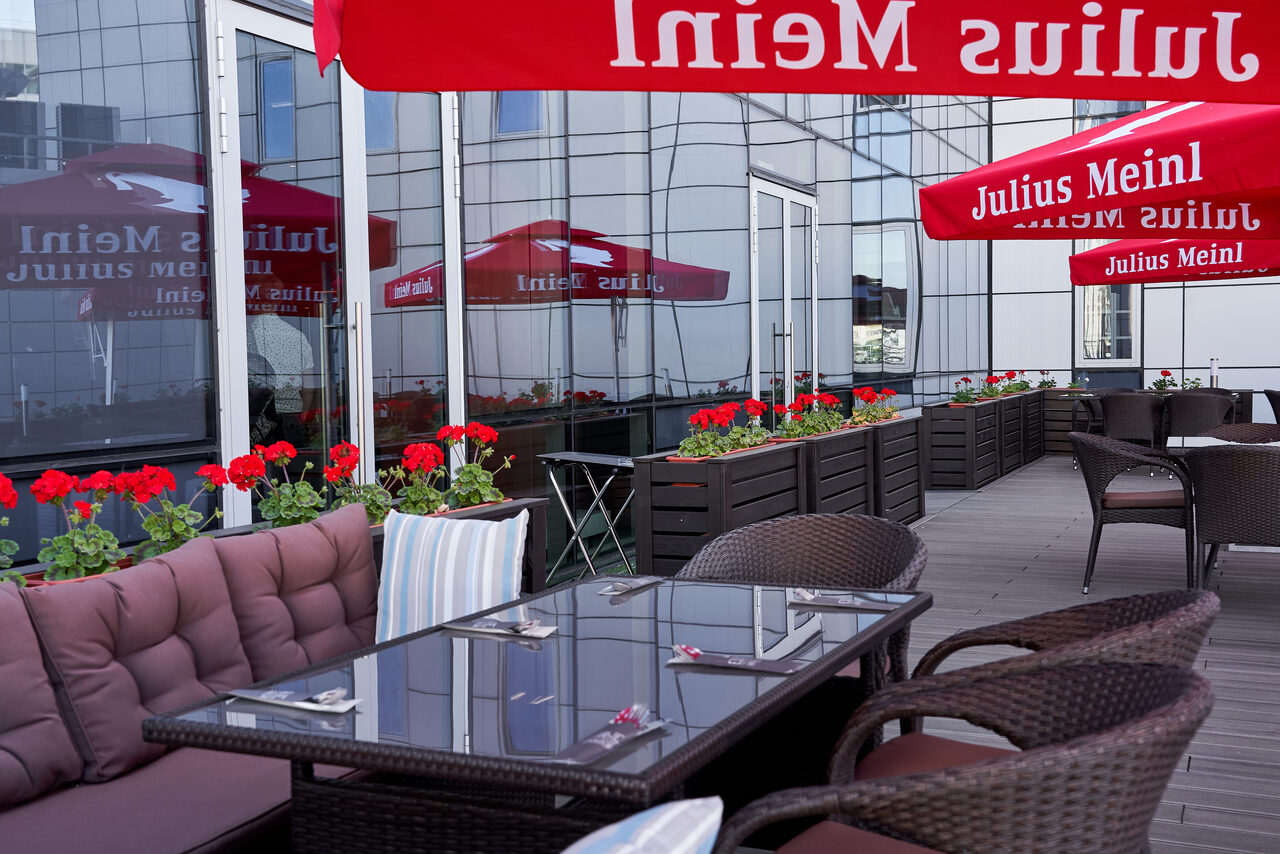 Арендовать Ресторан Ibis Kitchen(Ибис Кухня). Открытая терраса с плетёной мебелью: столы и кресла на деревянном настиле. Диван с коричневыми подушками, цветочные горшки с красными геранями. Красные зонты для тени. Стеклянные фасады здания на заднем плане. Надпись на зонтах.