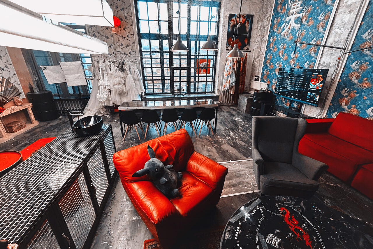 Арендовать Лофт Делюкс. Лофт-пространство с крупными окнами, деревянный пол, длинный стол с черными стульями, красное и серое кресла, мягкая игрушка, большая люстра, открытый гардероб с белыми платьями, телевизор, голубые обои с узором.