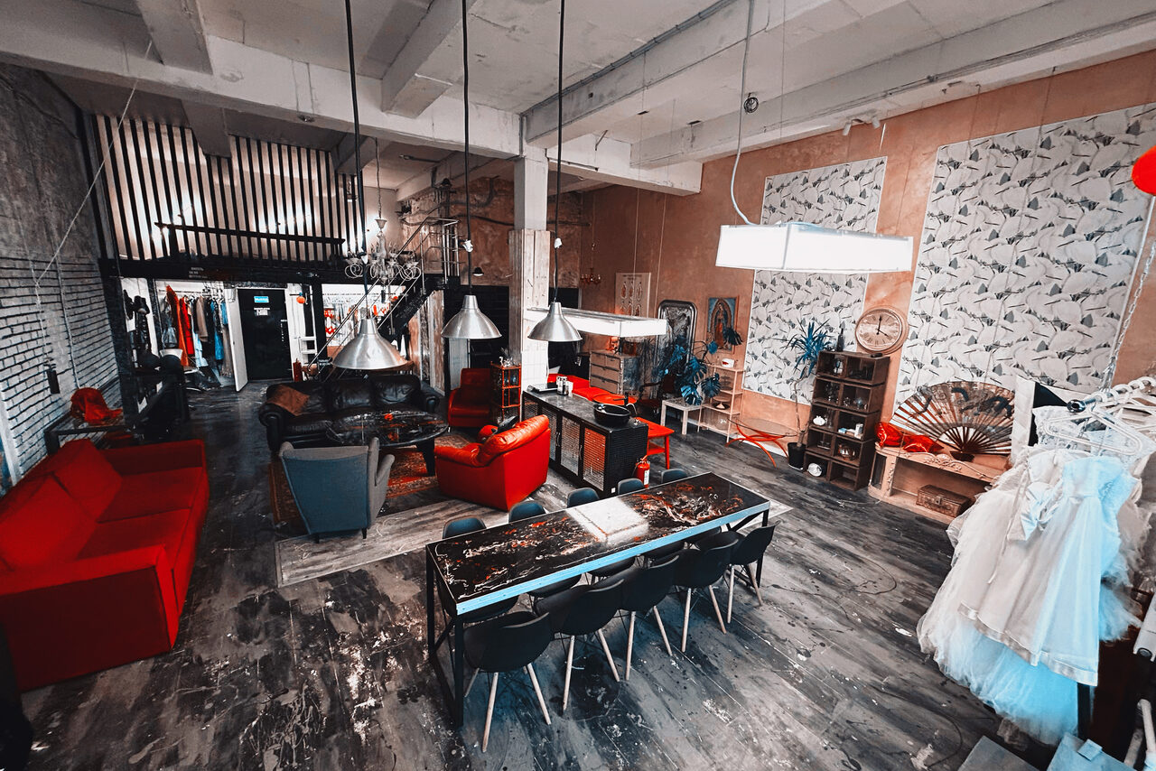 Арендовать Лофт Делюкс. Лофт-пространство с высокими потолками и белыми балками. Красный и серый диваны, длинный стол с черными стульями, промышленное освещение. Стена из кирпича, декор из растений и картин. Гардеробная зона и большое зеркало. Темный деревянный пол.