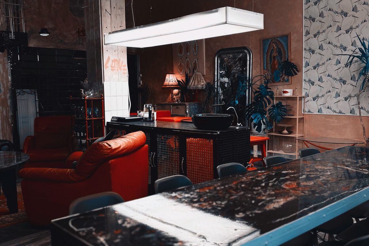 Арендовать Лофт Делюкс. Лофт-пространство с открытой планировкой. Красные кресла, длинный стол, подвесное освещение. Декор: картины, растения. Стены нейтральных тонов, часть с узором. Присутствуют кухонные элементы. На стене надпись "Love".