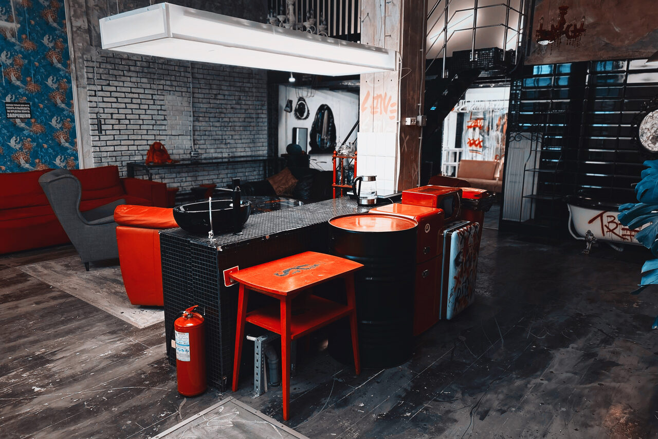 Арендовать Лофт Делюкс. Лофт-пространство с кирпичными стенами, серыми и красными элементами. Есть красные стулья, металлические бочки, комод и диваны. Светильники на потолке. На полу деревянное покрытие. Видны декоративные элементы и огнетушитель.