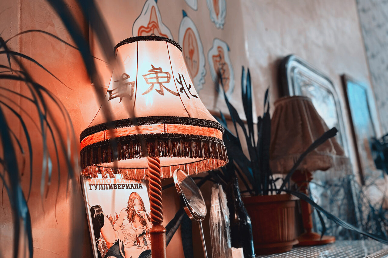 Арендовать Лофт Делюкс. Настольная лампа с абажуром, на котором есть символы, и бахромой, стоит рядом с растениями в горшках. На стене декор и рамки. На столе журнал с иллюстрацией. Освещение теплое, помещение оформлено в ретро-стиле.