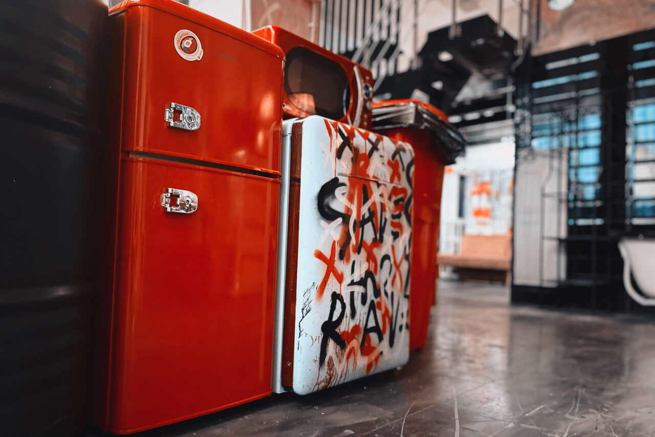 Арендовать Лофт Делюкс. Красный холодильник с рисунками граффити на передней панели. Пространство с бетонным полом и металлическими элементами, стеллажи на фоне. Смесь индустриальных и ярких акцентов.