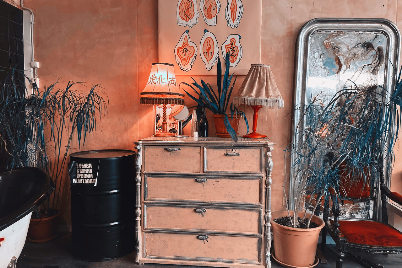 Арендовать Лофт Делюкс. Тип пространства: комната. Мебель: старинный комод, металлическая бочка. Освещение: лампы с абажурами. Материалы: дерево, металл. Декор: растения в горшках, большое зеркало. Цвета: бежевый, черный, красный. Состояние: винтажный стиль. Текст: надписи на бочке.