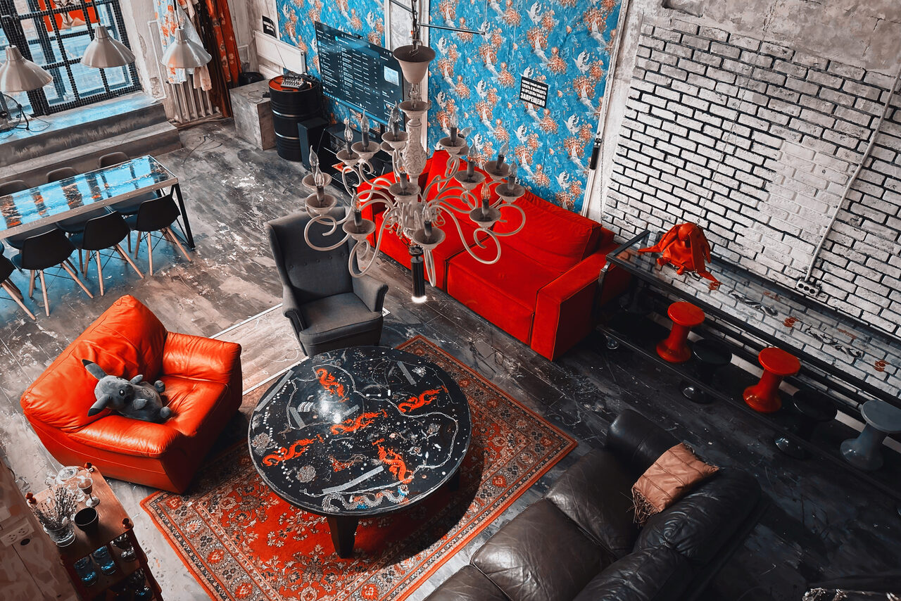 Арендовать Лофт Делюкс. Лофт-пространство с открытой планировкой, яркие цветные диваны и кресла, круглый стол с узором, кирпичная и цветочная обои на стенах, подвесные светильники, серый пол, ковёр с узором, высокие окна, барные стулья.