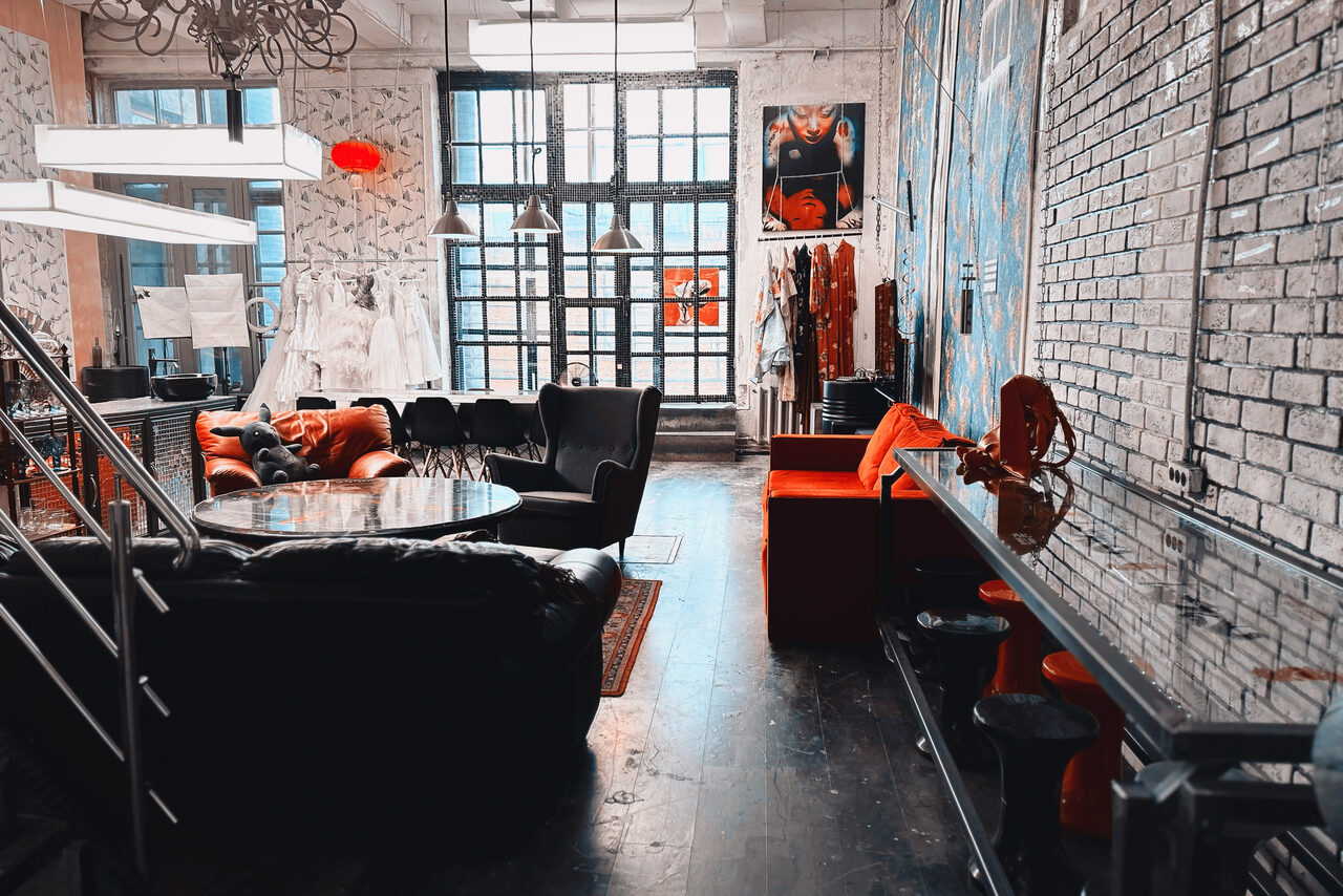 Арендовать Лофт Делюкс. Лофт-пространство с разноуровневыми потолками и большими окнами. Мебель: диван, кресла, столы. Стены из кирпича, светлые обои, яркие акценты. Освещение: люстры и подвесные светильники. На стенах постеры. Напольное покрытие — тёмное дерево.