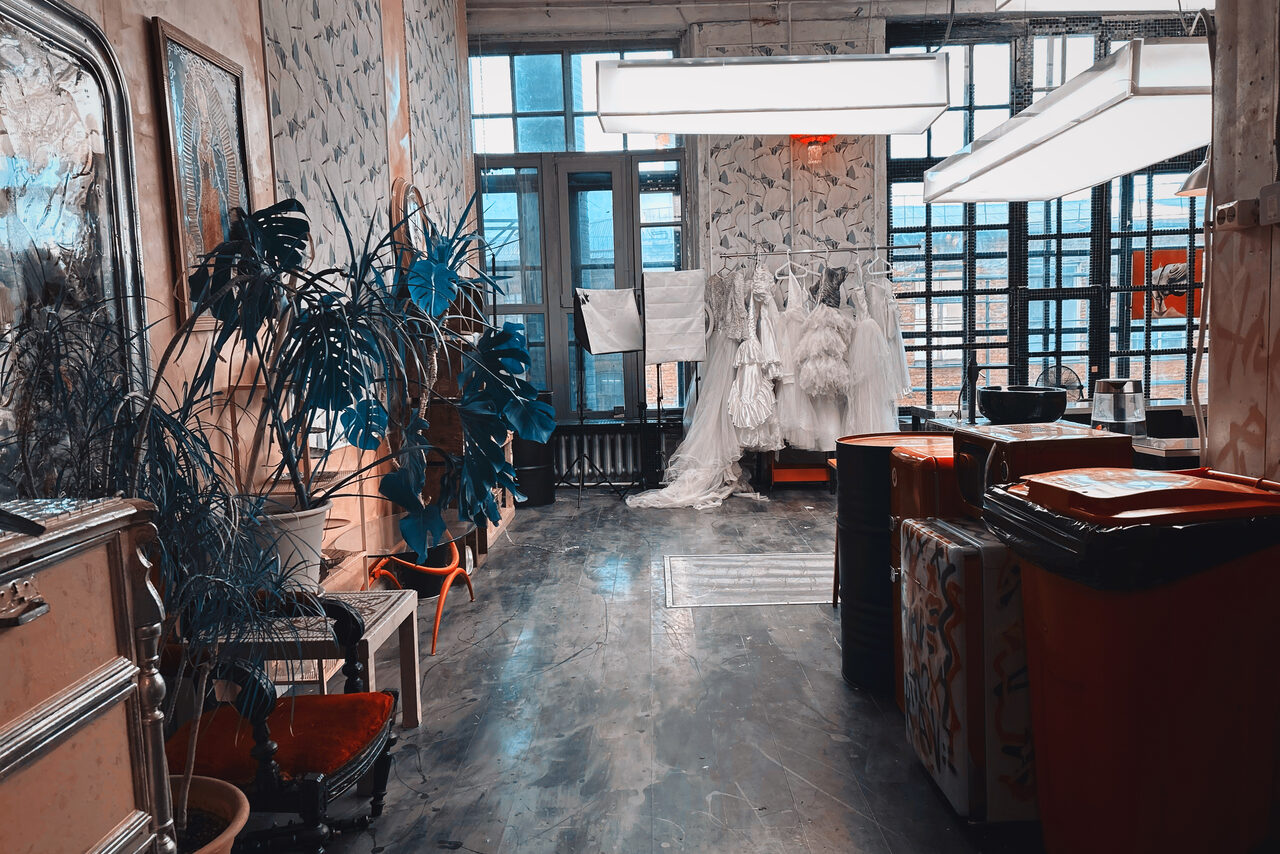 Арендовать Лофт Делюкс. Творческое пространство с большим окном, светлыми узорчатыми стенами, бетонным полом. В помещении несколько манекенов с одеждой, зеркальное трюмо, кресло, растения в горшках. Подвесные светильники прямоугольной формы, яркое освещение.