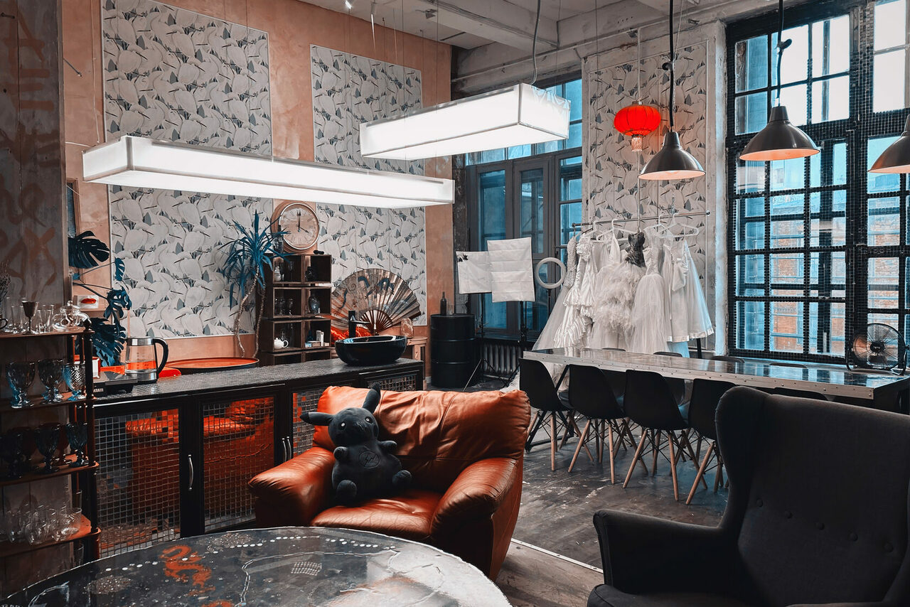 Арендовать Лофт Делюкс. Лофт-пространство с высокой мебелью. В центре коричневое кожаное кресло с мягкой игрушкой. Справа длинный стол с черными стульями, на вешалке висят белые наряды. На стенах растительный орнамент, крупные окна, подвесные светильники разного типа.