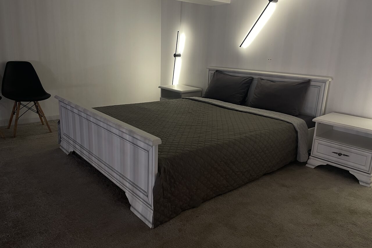 Арендовать Лофт Делюкс. Спальня с двуспальной кроватью с тёмным покрывалом и двумя подушками, двумя белыми прикроватными тумбочками. Справа черный стул. Вертикальное и наклонное настенное освещение. Светлые стены, ковролин на полу.