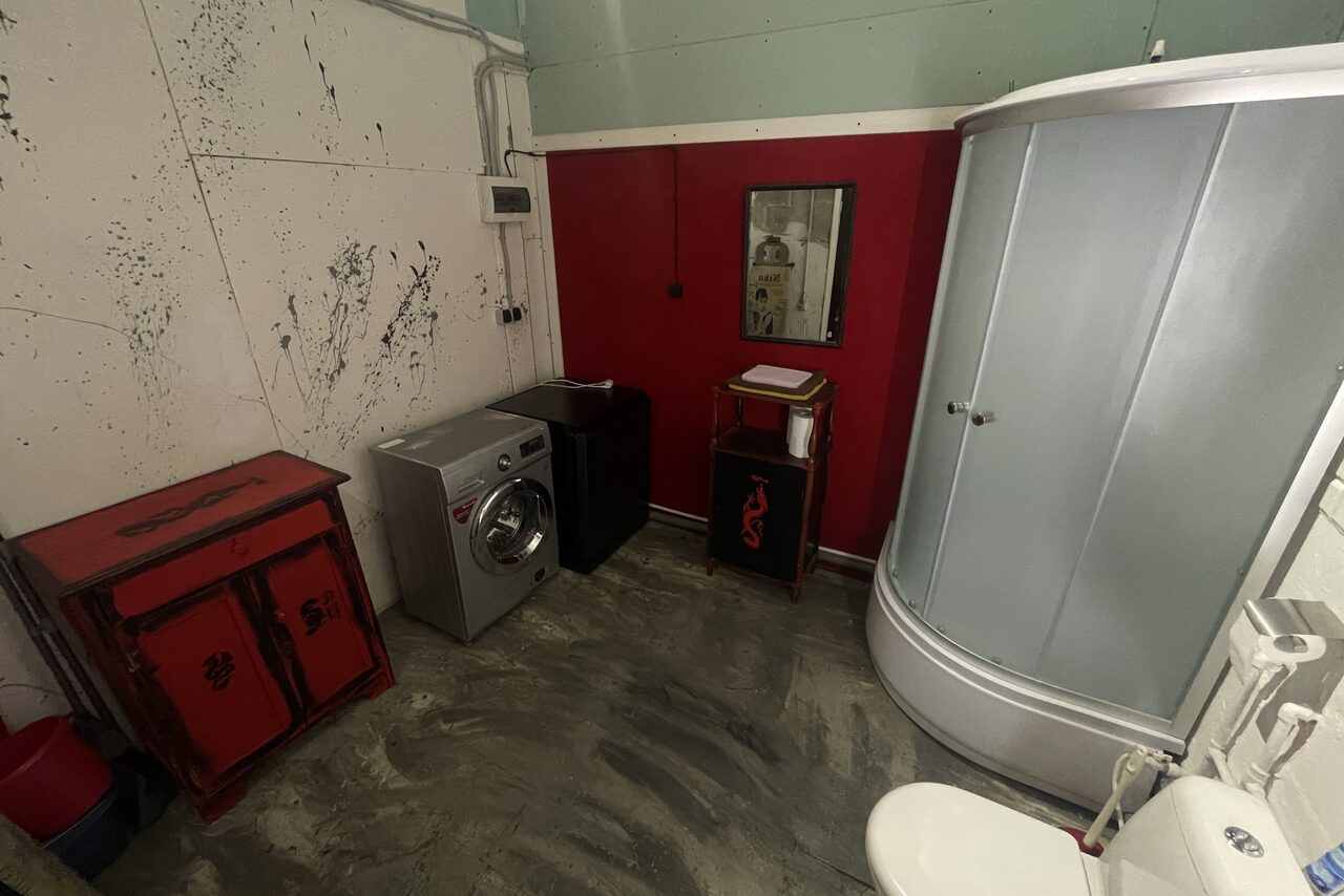 Арендовать Лофт Делюкс. Ванная комната с душевой кабиной, стиральной машиной и раковиной. На стенах красные панели и отделка с черными узорами. Напольное покрытие серого цвета. Присутствует зеркало, шкафчики и полка. Освещение потолочное.