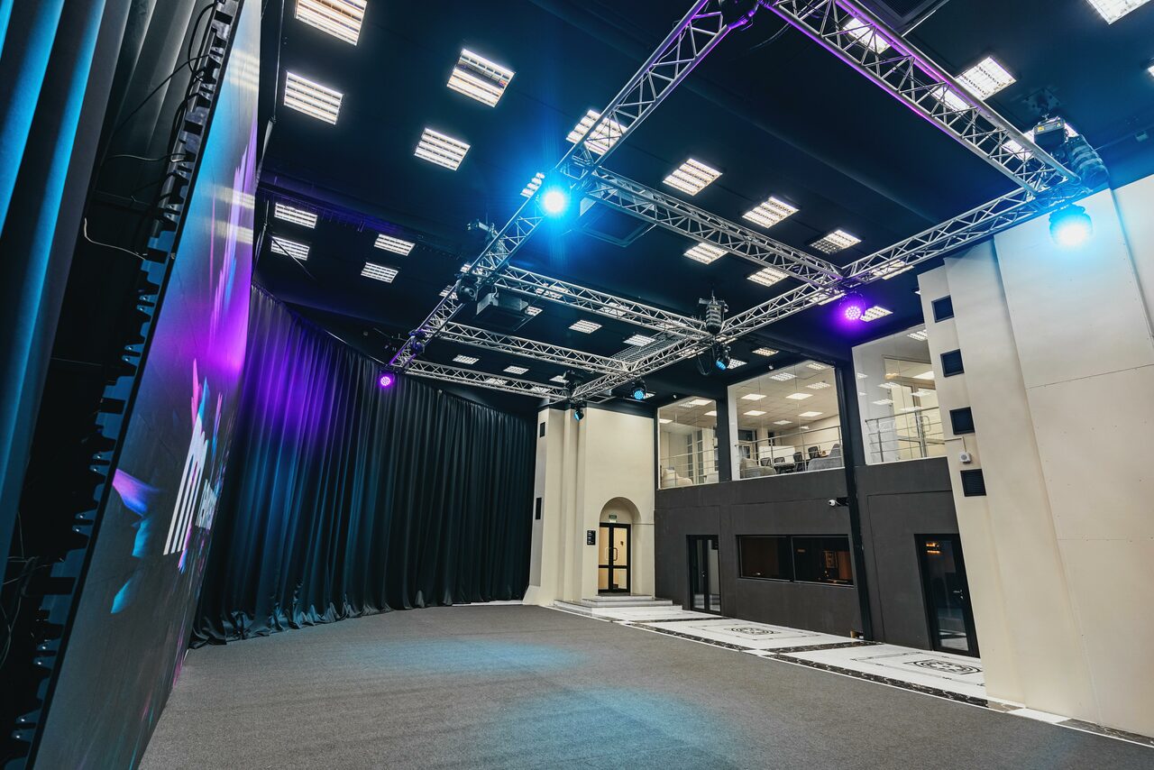 Арендовать New Media (Нью Медиа) продакшн. Павильон с большим LED-экраном. Просторное помещение с высокими потолками и сценой. На стене большой экран. По периметру установлены световые конструкции с прожекторами. Пол ковровый, стены светлые с черными акцентами. Сбоку два уровня с окнами и дверями.