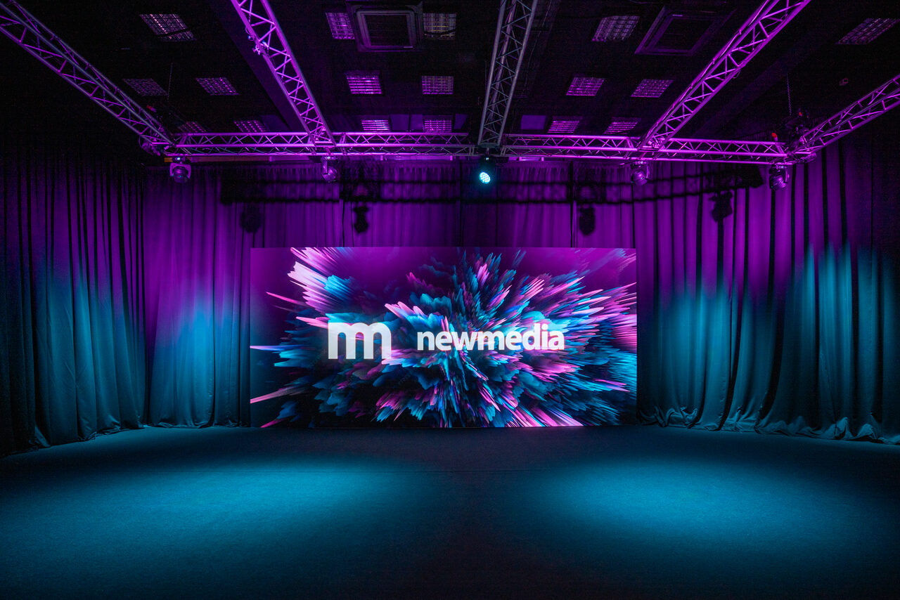 Арендовать New Media (Нью Медиа) продакшн. Павильон с большим LED-экраном. Просторное помещение с черными тканевыми драпировками по периметру и сценой. В центре яркий экран с абстрактным дизайном и текстом "newmedia". Потолок с подвесной конструкцией и фиолетовым освещением. Пол темного цвета.