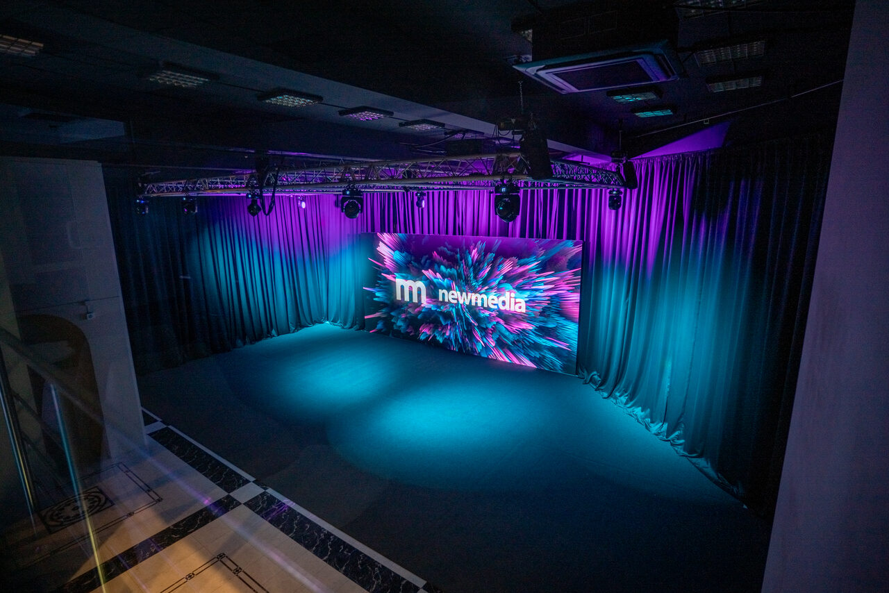 Арендовать New Media (Нью Медиа) продакшн. Павильон с большим LED-экраном. Сценическое пространство с темными шторами и экраном, на котором виден текст "newmedia". Освещение фиолетового и синего оттенков, потолок с квадратными светильниками. Помещение в хорошем состоянии.