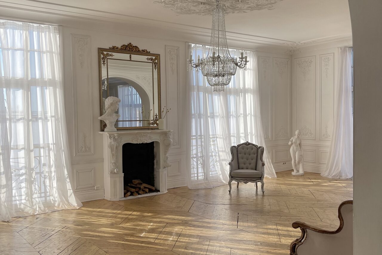 Арендовать Арт-Плейс Фактура. Просторный лофт в классическом стиле. Светлая комната с высокими потолками, паркетным полом. У стены декоративный камин с зеркалом и бюстом. На потолке люстра. У окна серое кресло. Боковые стены украшены лепниной, имеются плинтусы. Шторы из легкой ткани.