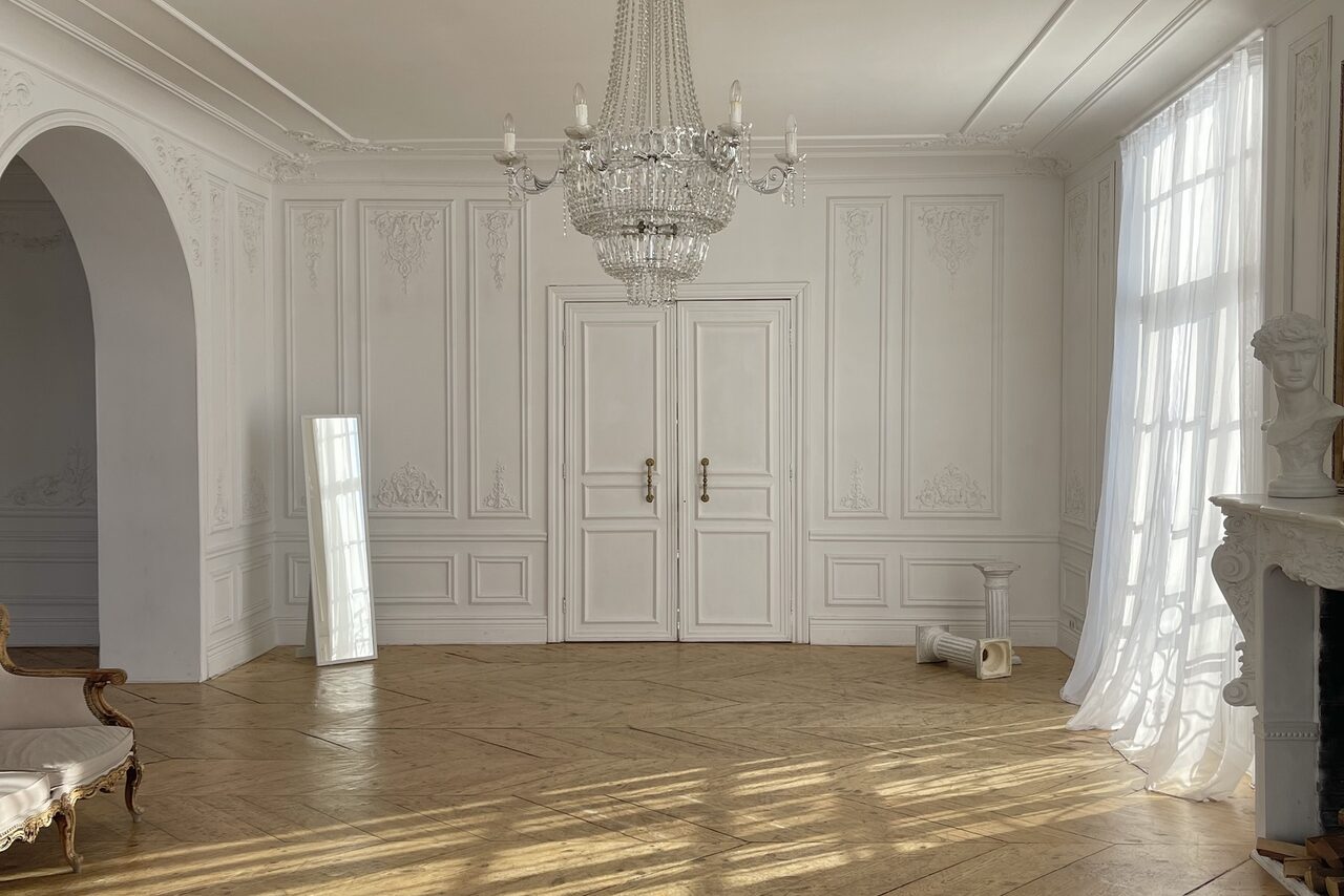 Арендовать Арт-Плейс Фактура. Просторный лофт в классическом стиле. Просторная светлая комната с белыми стенами и высокими потолками. На полу паркет, крупная люстра, арка, белые двустворчатые двери. У стены стоит зеркало, у окна бюст, рядом белая колонка. Длинные полупрозрачные шторы закрывают большое окно.