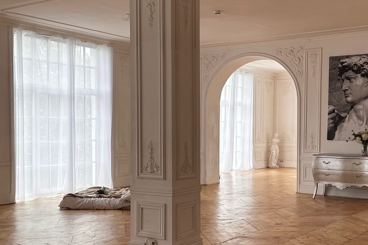 Арендовать Арт-Плейс Фактура. Просторный лофт в классическом стиле. Просторное светлое помещение с арочными проемами и высокими французскими окнами. На полу паркет, стены украшены молдингами. Подушка на полу, белая мебель, черно-белое изображение бюста. Растение в вазе на комоде.