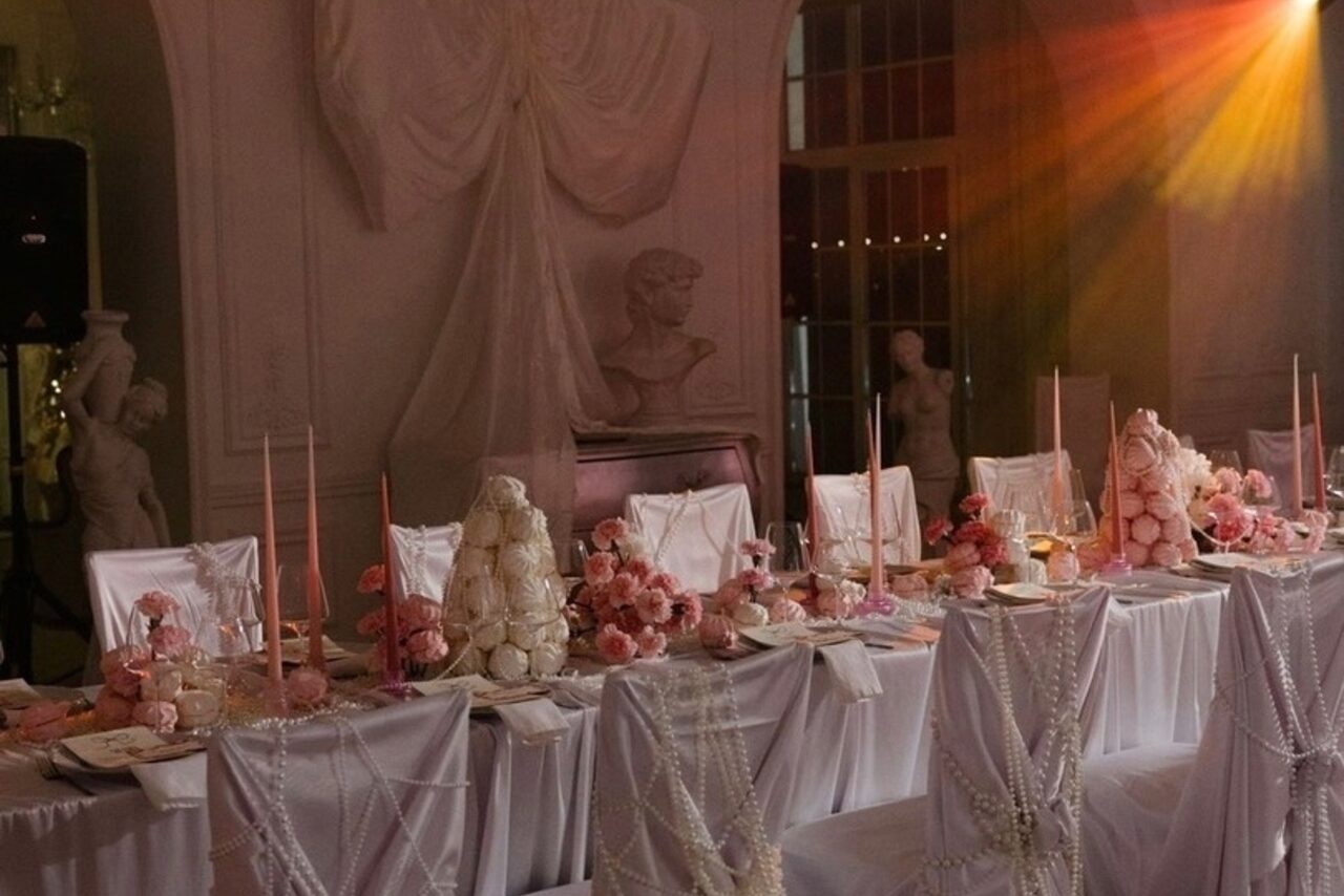 Арендовать Арт-Плейс Фактура. Просторный лофт в классическом стиле. Банкетный зал с длинным накрытым столом, белые скатерти и чехлы на стульях, декор из розовых цветов и свечей, скульптуры на стенах, мягкое освещение теплых тонов, на стене белый архитектурный декор и бюст.