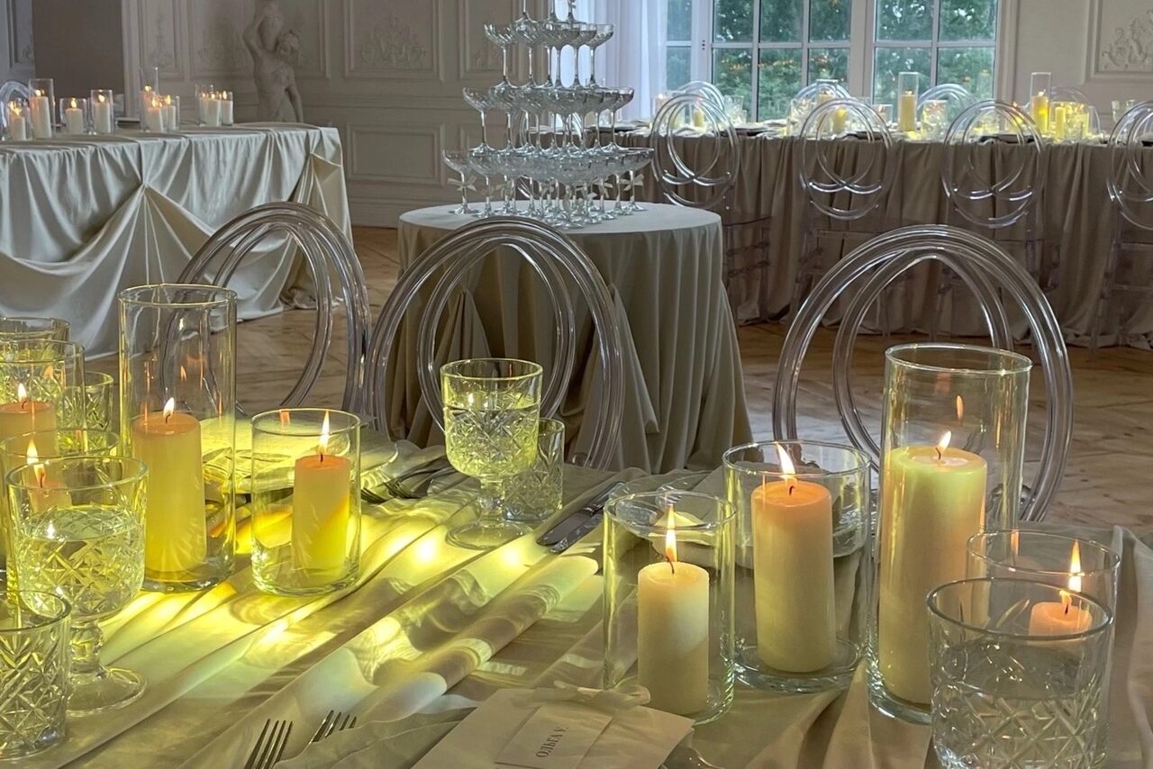 Арендовать Арт-Плейс Фактура. Просторный лофт в классическом стиле. Банкетный зал с сервированными столами и стеклянными стульями. На столах свечи в подсвечниках, салфетки, бокалы. Декор из тканей. На заднем плане высокий пирамидальный стеклянный декор. Светлое наклонное освещение из окна. Пол паркетный.