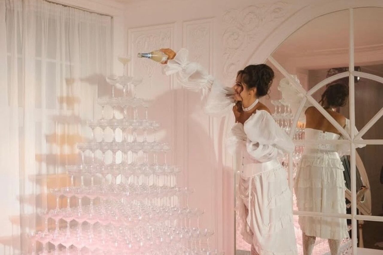 Арендовать Арт-Плейс Фактура. Просторный лофт в классическом стиле. Женщина в белом платье наливает напиток из бутылки в бокалы на стеклянной пирамиде. Светлая комната с декоративными панелями и большим зеркалом. Теплое освещение, белые стены, светлый текстиль.