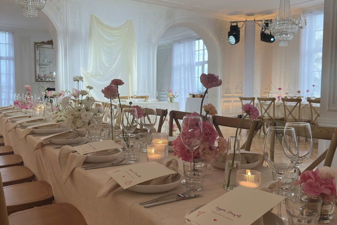 Арендовать Арт-Плейс Фактура. Просторный лофт в классическом стиле. Просторная банкетная комната с длинным столом, накрытым светлой скатертью, украшенным розовыми и белыми цветами. Деревянные стулья, свечи, бокалы, карточки на тарелках. Белые стены, арочные окна с белыми шторами, хрустальная люстра на потолке, светлое напольное покрытие.