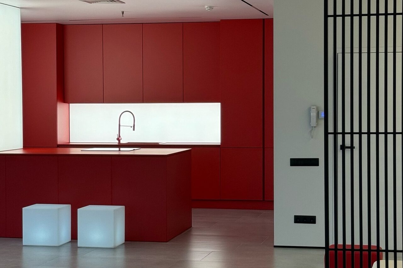 Арендовать Арт-Плейс Фактура. Современный лофт с видом на весь город. Кухонное пространство с красными шкафами и островом, встроенная мойка с изливом, светлое фоновое освещение, два белых кубических сиденья, светлый деревянный пол, черные решётки перегородки и настенный домофон.