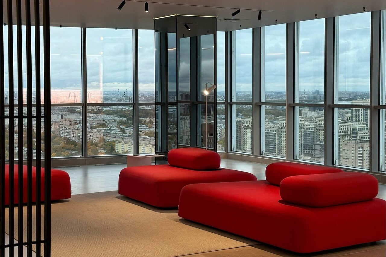 Арендовать Арт-Плейс Фактура. Современный лофт с видом на весь город. Современное пространство с панорамными окнами, открывающими вид на город. В центре расположены красные мягкие диваны. Потолочное освещение из множества направленных светильников. Стены и пол выполнены из светлых материалов.