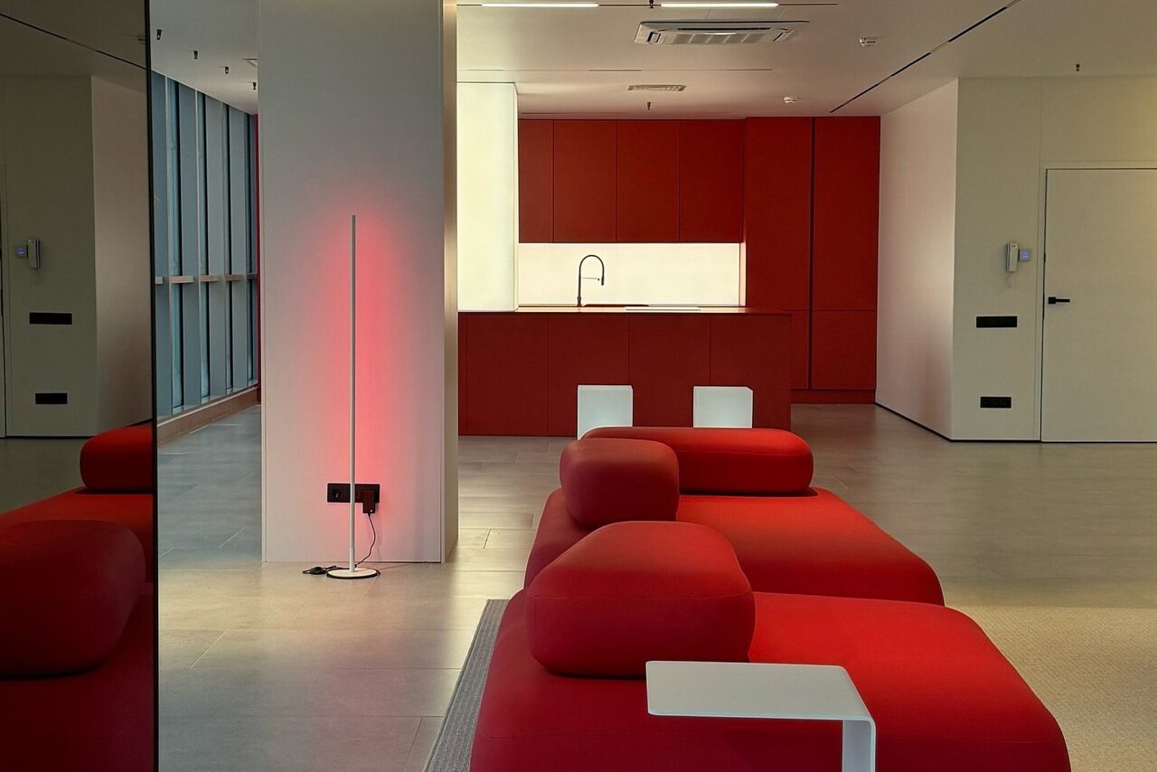 Арендовать Арт-Плейс Фактура. Современный лофт с видом на весь город. Современное пространство с открытой планировкой, красной кухней и красными модульными диванами. Встроенный светильник, белый столик. Большие окна, деревянный пол, светло-бежевые стены. Зеркало на стене, белая дверь.