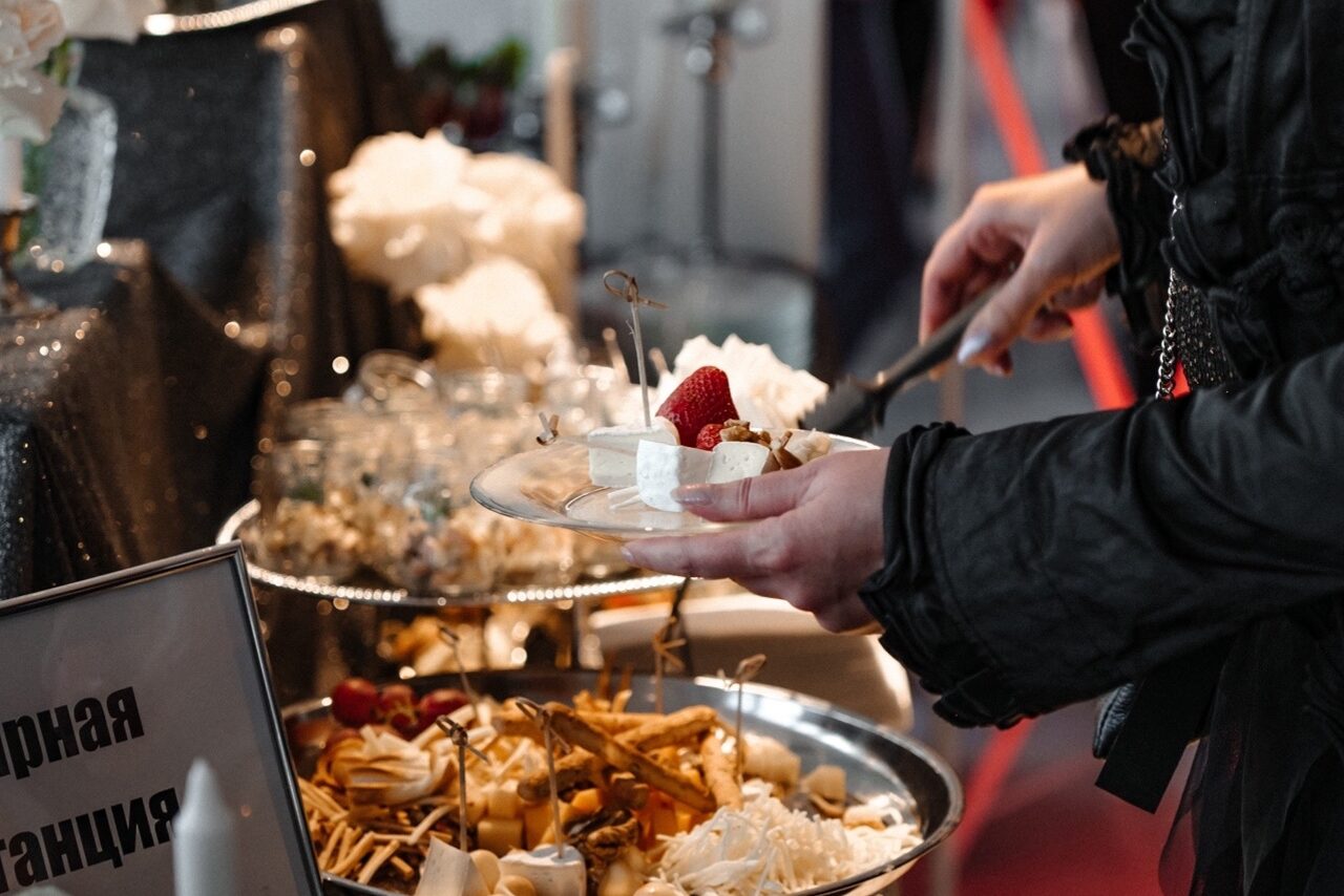 Арендовать Арт-Плейс Фактура. Современный лофт с видом на весь город. Человек держит тарелку с десертами, включая кусочки торта и клубнику. На столе рядом стоит большой поднос с различными закусками и украшениями. На табличке видна частичная надпись. Освещение мягкое, обстановка праздничная.