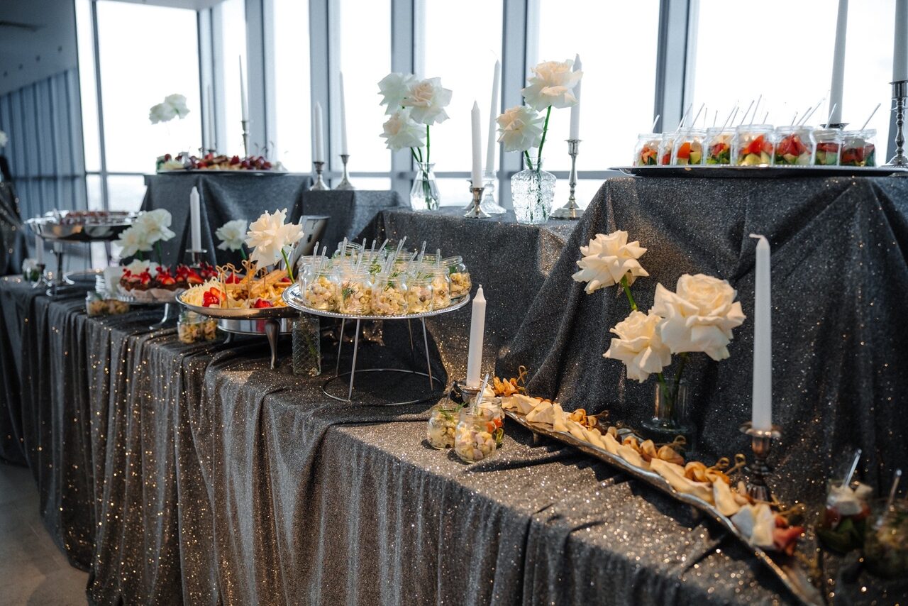 Арендовать Арт-Плейс Фактура. Современный лофт с видом на весь город. Стол с серой блестящей скатертью, на котором расставлены вазы с белыми цветами, высокие белые свечи и блюда с канапе и десертами. Стены с крупными окнами пропускающими много света.