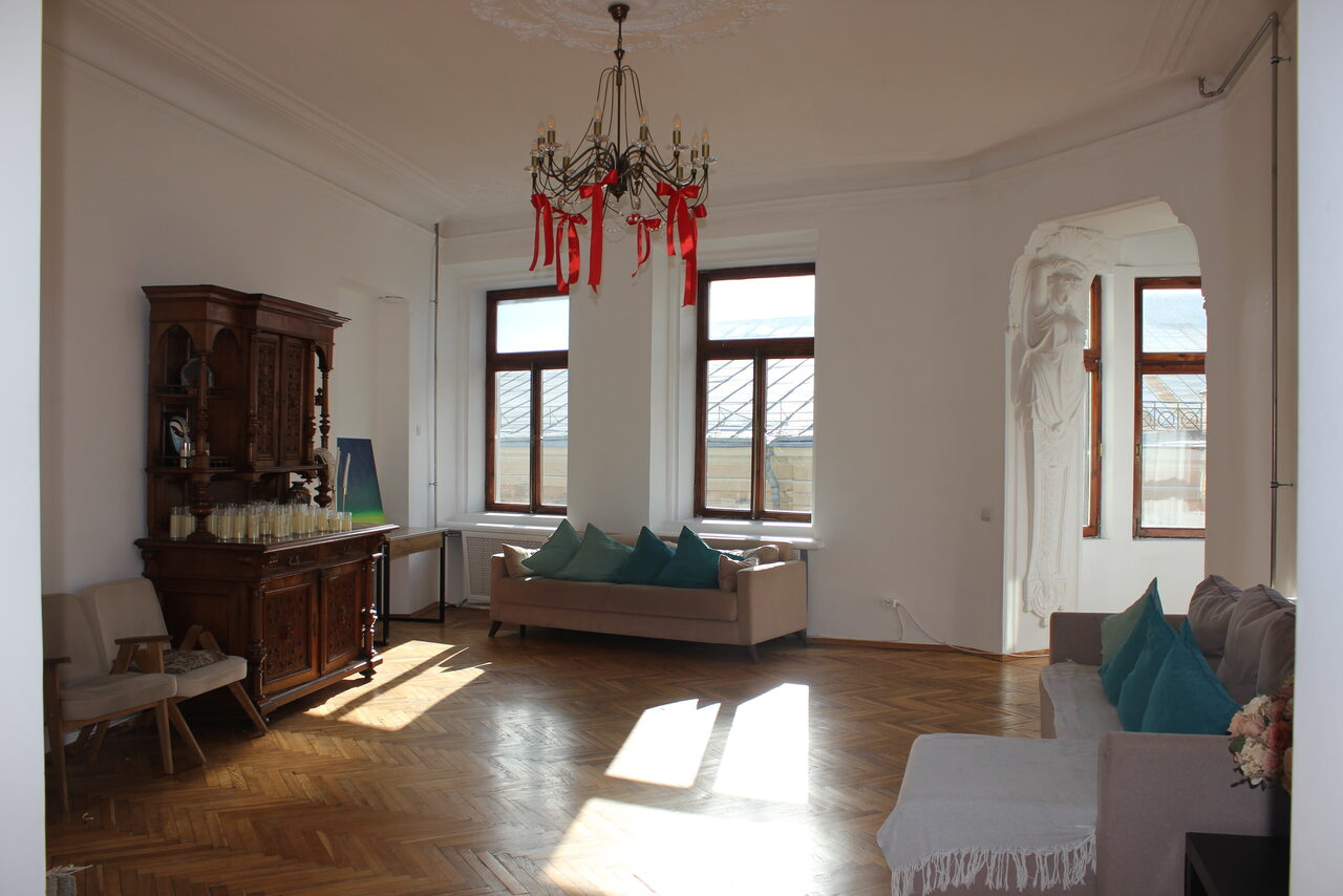 Арендовать Cassiopea (Кассиопея). Светлая гостиная с деревянным паркетом, два дивана с подушками, антикварный шкаф, стулья. Большие окна, люстра с красными элементами. Белые стены, скульптурная ниша. Мягкий дневной свет.